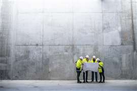 A group of four engineers in hard hats and yellow hi-vis jackets examine plans against the backdrop of a concrete wall
