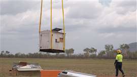 Large box being lifted by helicopeter (out of sight) with worker looking on