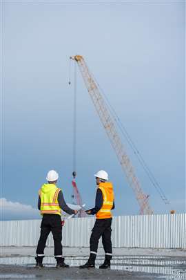 Two construction engineers wearing safety vests and helmets stand on a wet site, discussing with documents in hand while a large crane operates in the background under a clear sky.